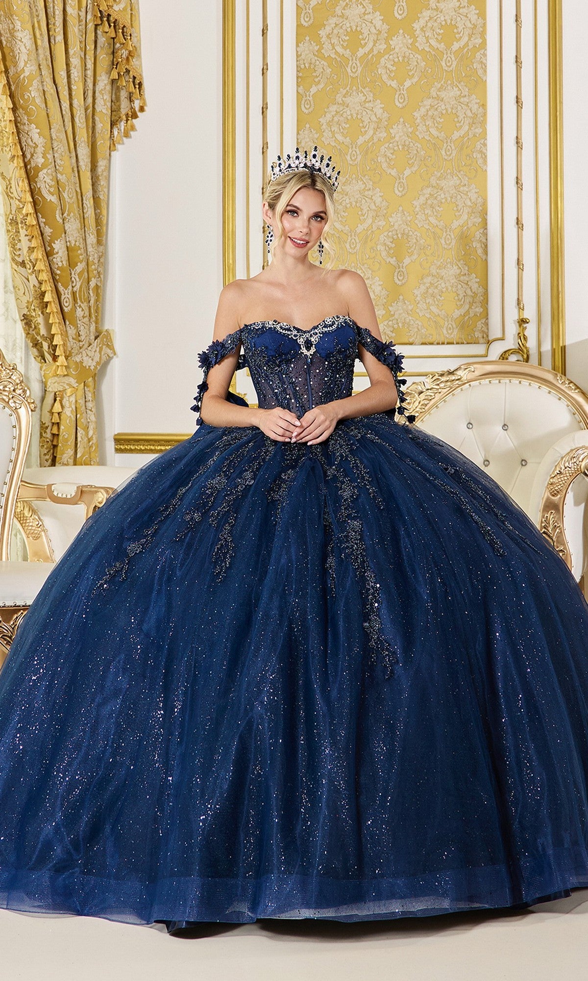 A woman wearing the Quinceanera Dress 5083 by Dancing Queen in navy and a tiara poses gracefully in an ornate room with gold and cream decor, standing in front of an elegant sofa.